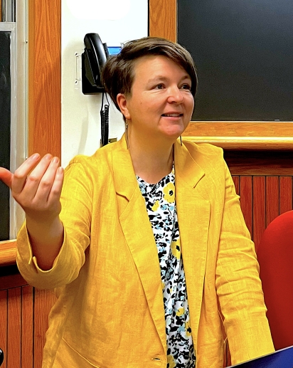 Centered in the image is a person wearing a bright yellow linen suitcoat and a dappled black and yellow shirt. She is looking off camera to the right, smiling and gesturing. Behind her is a blackboard and a classroom phone.