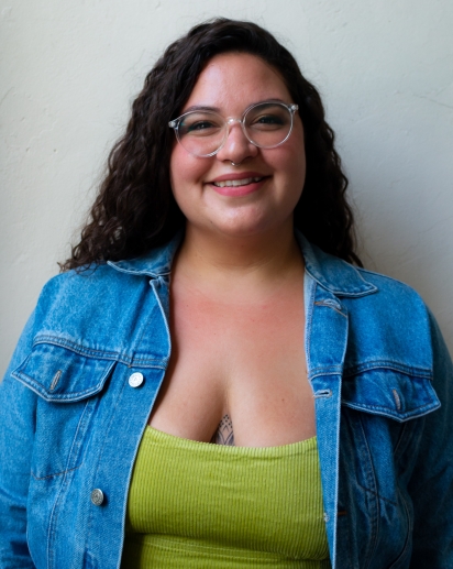 Photo of Margarita Hernandez. Margarita is wearing a green dress with a jean jacket. They have long curly hair and transparent glasses. They have beige skin with red and pink undertones. They are smiling. 