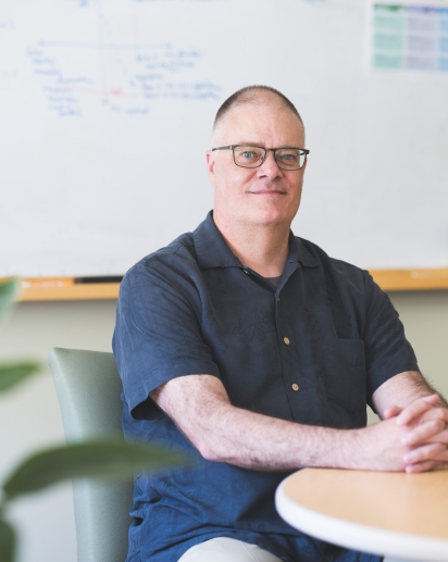 A photograph of Scott Pauls sitting at a table in front of a whiteboard.  Image Credit: Katherine Lenhart, 2025 